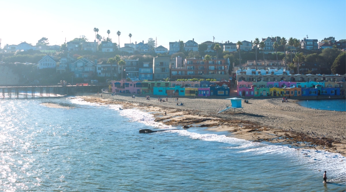 A top view capturing Capitola Beach in Santa Cruz. The image features the ocean on the left and the beach on the right, bordered by a line of colored houses in the background. The photograph provides an aerial perspective of the coastal setting, highlighting the expanse of the beach and the adjacent ocean.