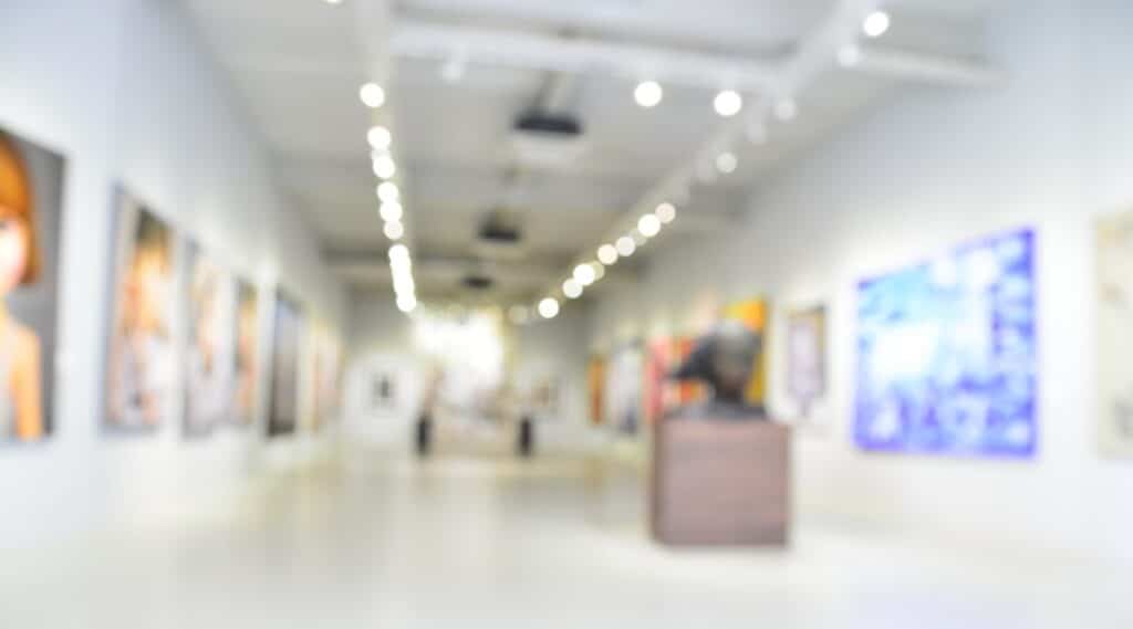 Blurred image of the lobby of a modern art center. The hall is bright, with white walls, with warm white lighting. There are paintings on the walls, and sculptures are displayed in the middle of the hall.