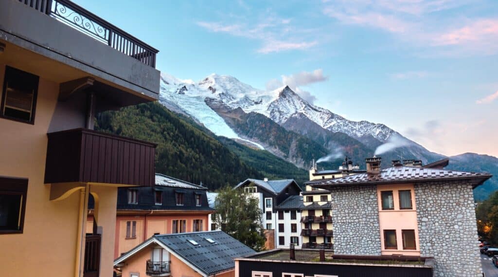 A photograph capturing the autumn mountain landscape in Chamonix, France. In the foreground, various city houses of different colors and architectural styles are visible. The background features the Mont Blanc massif, characterized by snow-capped mountains and rocky formations.