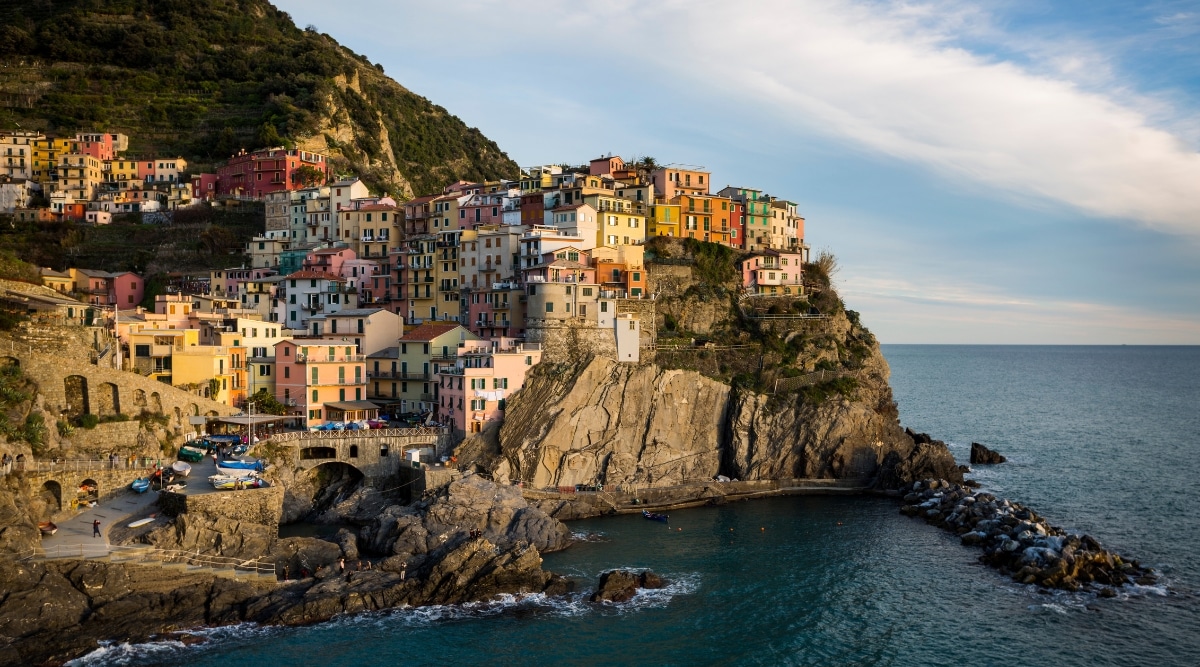 A panoramic view capturing the coastal landscape of Riomaggiore in Cinque Terre, featuring the sea and the vibrant facades of houses along the shoreline. The image showcases the characteristic architecture of the region, with multicolored houses lining the waterfront. The sea is visible in the foreground.
