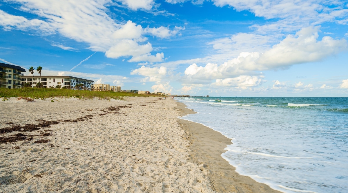A panoramic view capturing Cocoa Beach, Florida, with the beach and residential houses visible on the left side and the expanse of the ocean on the right. The beach stretches across the frame without any discernible presence of people. The coastal scene is under a sky with a few clouds.