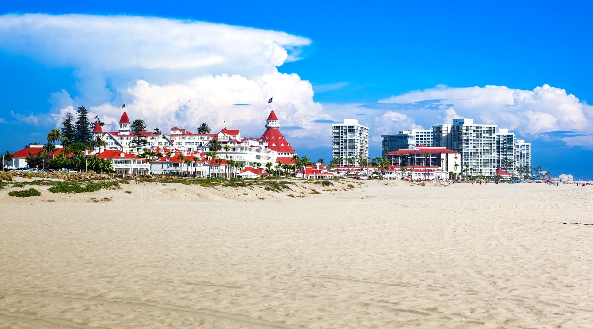 The image depicts a view of Coronado Beach Island in San Diego. In the foreground, there is a stretch of sand, and in the background stands a notable hotel featuring a red roof. The sky is characterized by a bright atmosphere with large white clouds.