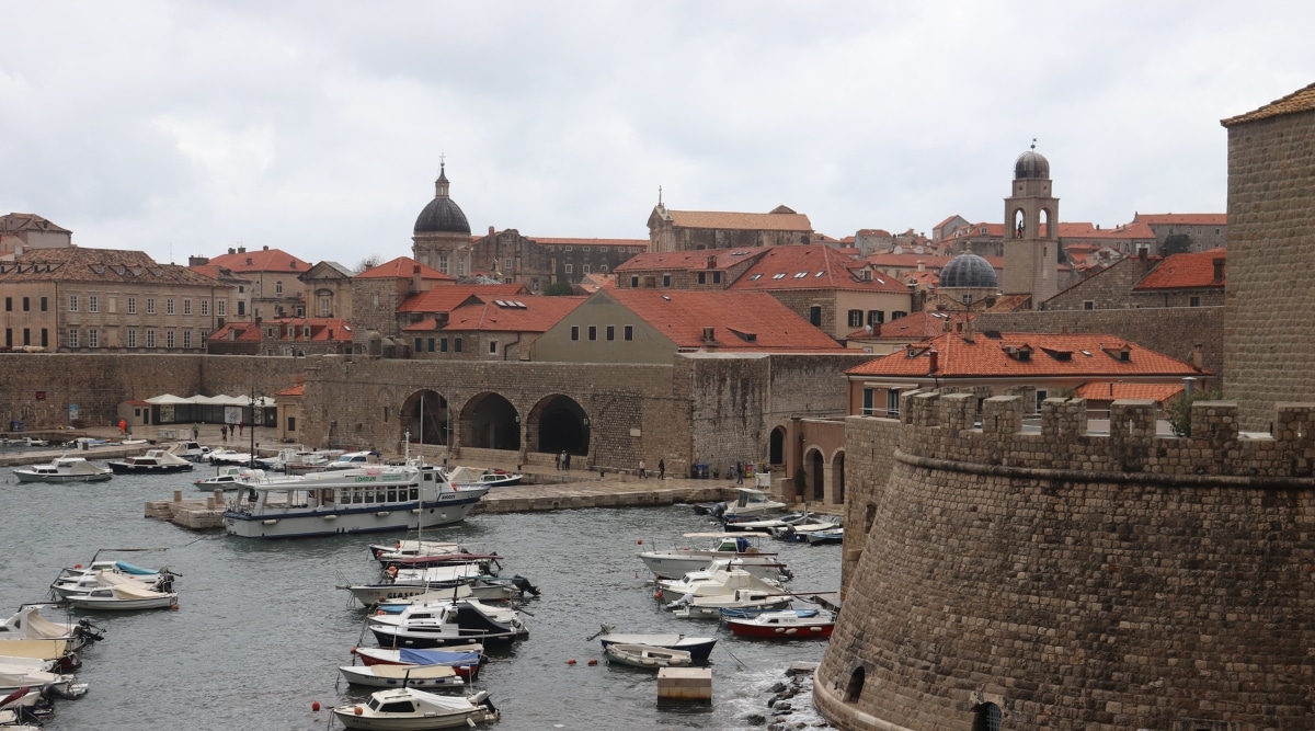 A view of Dubrovnik, Croatia, depicted in the image with a prominent pier featuring various boats and ships. The cityscape reveals buildings with characteristic red roofs against an overcast sky.