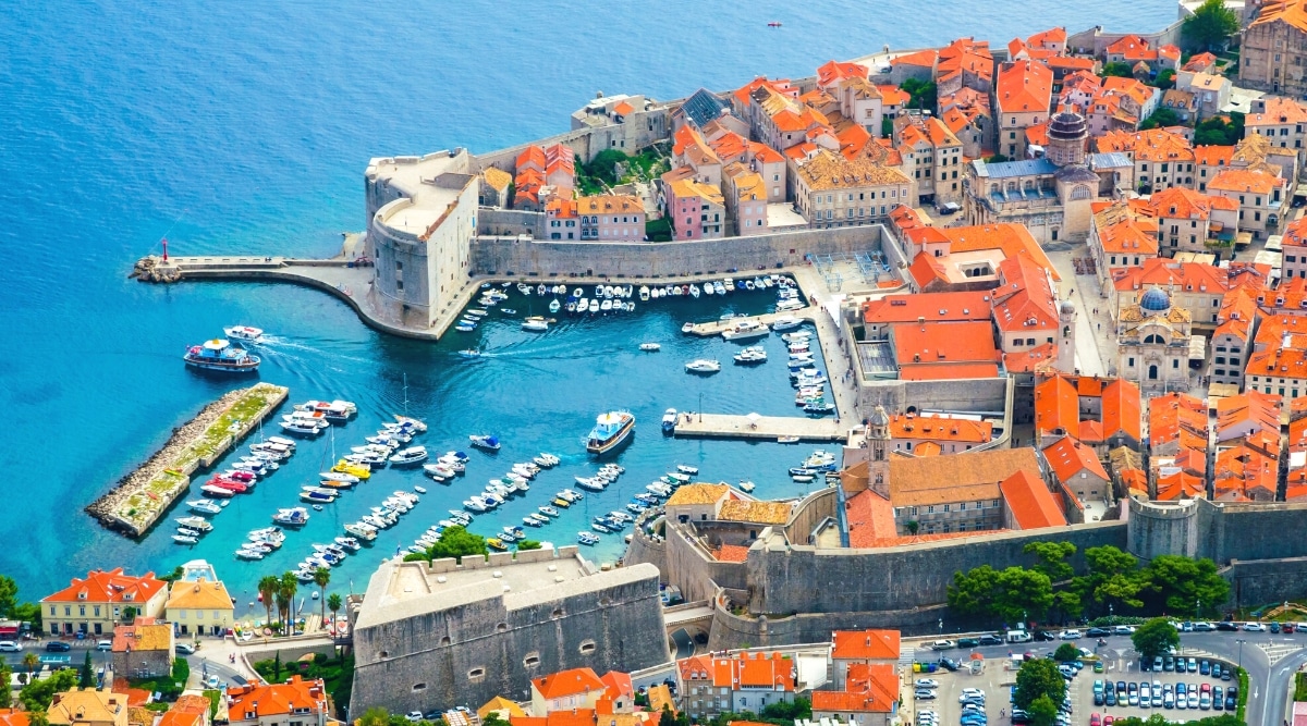 An aerial photograph capturing the cityscape of Dubrovnik, Croatia. The image features the city's architecture and coastal area, including a pier with a substantial number of yachts moored along it. The city is characterized by its distinctive red-roofed buildings, extending to the coastline.