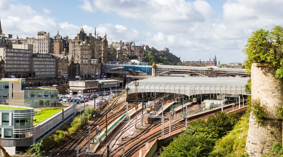 An aerial view capturing Edinburgh Waverley against a sunny backdrop with scattered clouds. The image showcases the train station's infrastructure and platforms, extending towards the cityscape in the background.