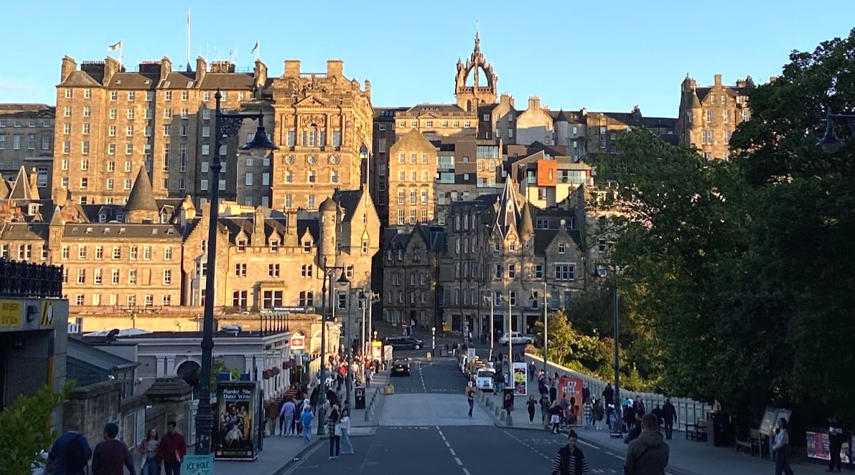 The image captures Edinburgh's Old Town, featuring a straight street in the foreground bustling with pedestrian activity. The background showcases ancient buildings, approximately 10-15 floors high, characterized by historic architectural design.