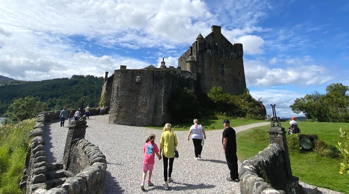 The image captures Eilean Donan Castle, with visitors exploring the historical site. The weather is favorable, marked by light clouds, and the surroundings feature abundant greenery. The castle's architectural features are visible against the backdrop of the scenic landscape.