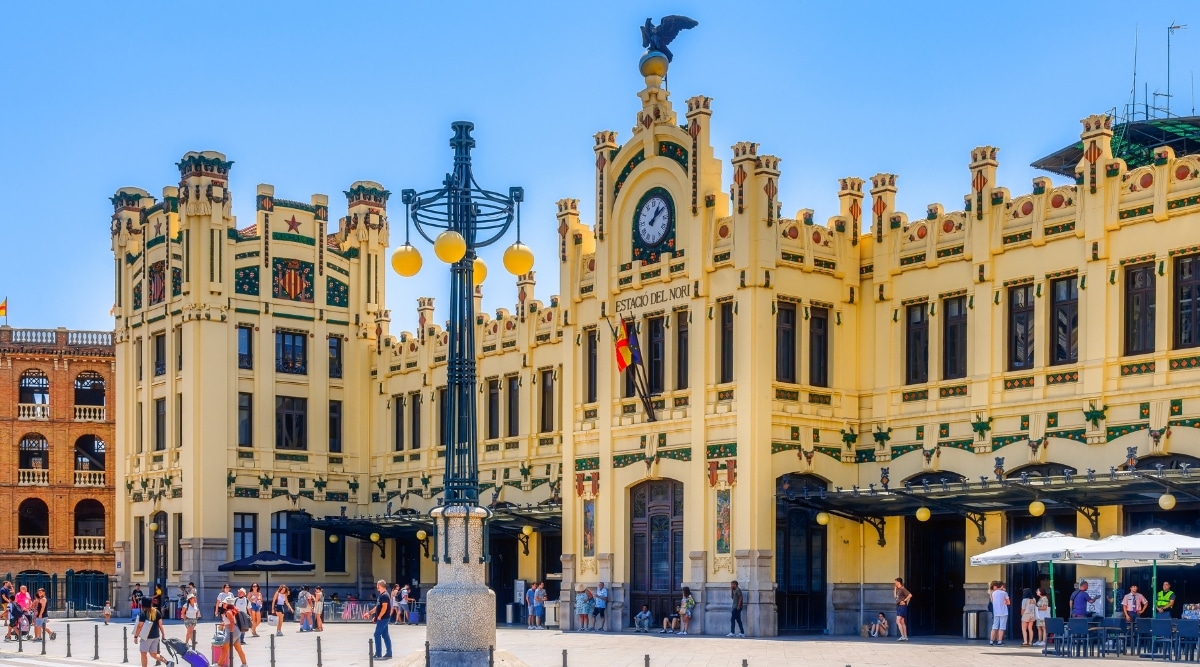 A view from ground level capturing Estacion Del Norte in Valencia, Spain. The image features a yellow building with a centrally positioned clock. Several dozen people are visible around the station. 