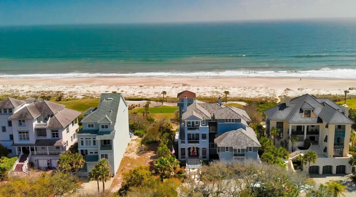 A view capturing Fernandina Beach, Florida, featuring four prominent houses in the foreground amid abundant greenery. The houses are distinctively visible, and the scene extends to showcase an empty beach with the ocean in the background. 