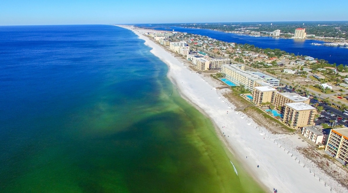 An aerial view capturing Fort Walton Beach, Florida, with a clear sky overhead. The image reveals a balanced composition with the cityscape on the right, featuring multi-story buildings, and the ocean on the left. The beachfront is prominent, displaying a combination of natural landscapes and urban development.