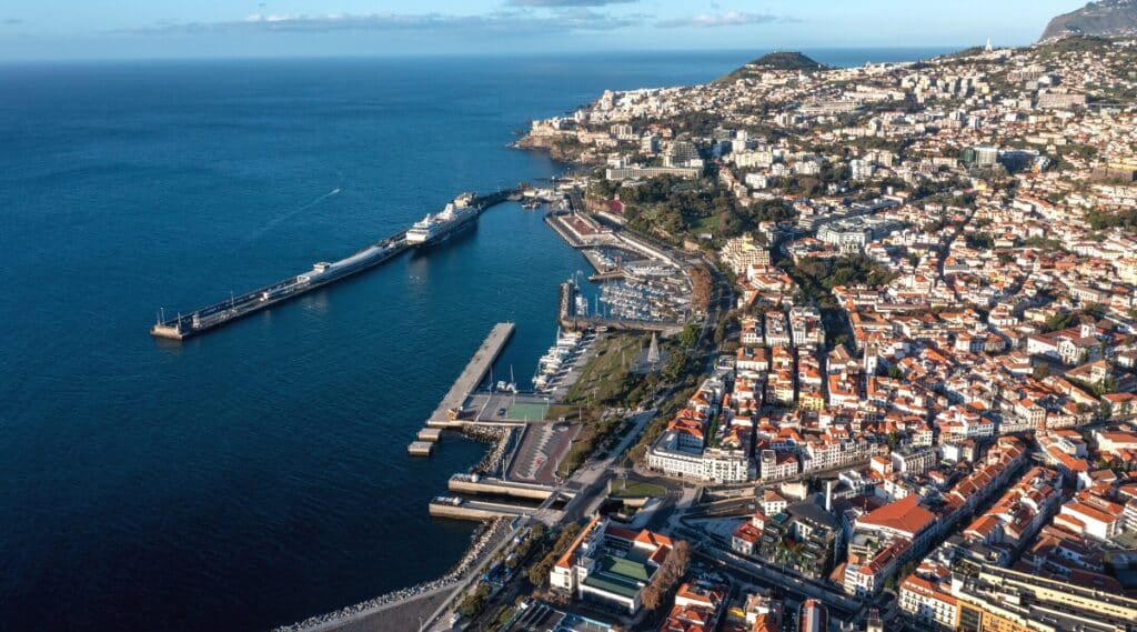 A drone view capturing Funchal, Madeira, on a clear day. The image features the cityscape characterized by orange rooftops, showcasing the architectural layout of the urban area. A pier is visible, hosting both large and small ships and boats.
