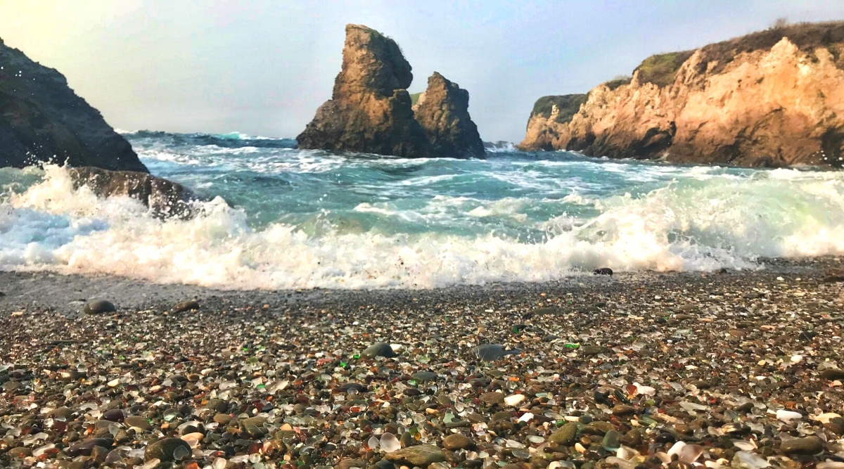 A view of Glass Beach in Fort Bragg, featuring a foreground coastline adorned with smoothed colored glass fragments, a result of natural weathering. The ocean extends in front of the beach, providing a maritime backdrop, while rocks and hills form the background terrain.