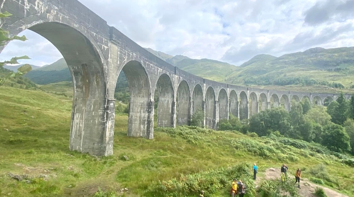 A photograph capturing the Harry Potter Bridge from a lower perspective, set against an overcast sky. The image showcases the iconic bridge's architectural details and its surroundings. Abundant greenery surrounds the bridge, providing a natural backdrop. Four tourists are observed exploring the area.