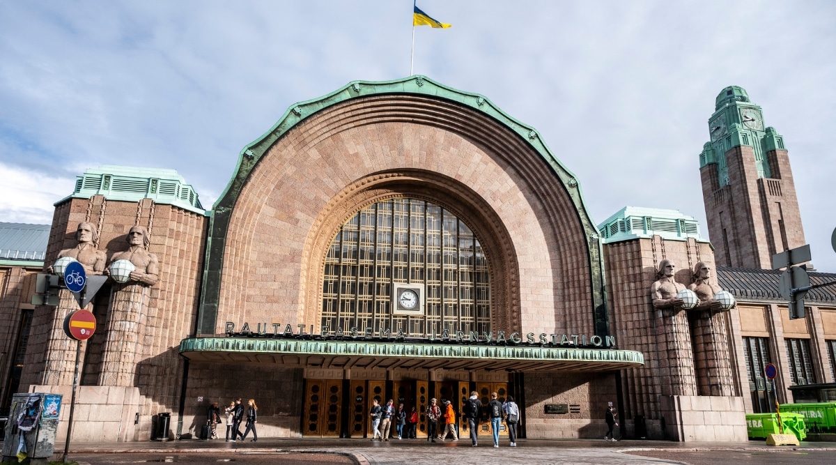 A ground-level view capturing Helsinki Central in Finland. The image features the train station building with its distinct architectural elements. Several pedestrians are visible in the surroundings. The station building exhibits a classic architectural style. 