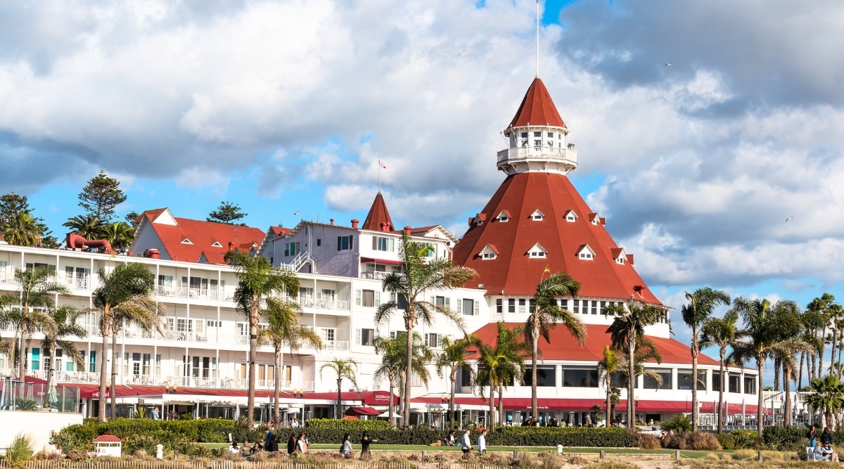 A clear day view capturing Hotel Del Coronado, a high-rise structure with a distinctive red roof. The image showcases the hotel's architectural features against a backdrop of large clouds in the sky. The surrounding landscape includes numerous palm trees.