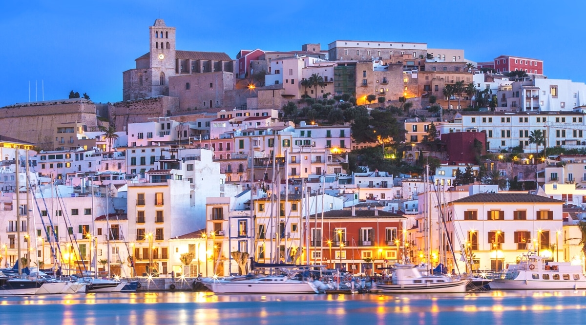 A drone view captured from the sea showcasing the city of Ibiza, Spain during the evening. Numerous buildings ascend the hill, and boats are visible moored in the foreground. The cityscape is illuminated by street lights, defining the landscape. 