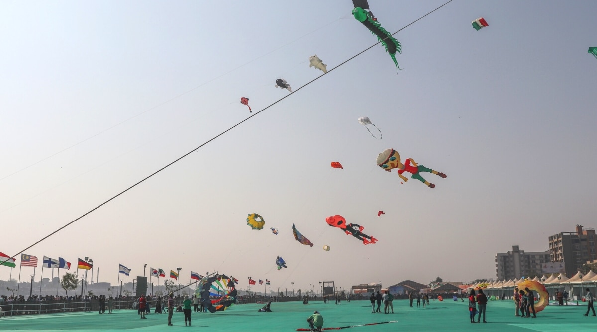 A photograph capturing the scene at the International Kite Festival in Gujarat, India. The image features a sky filled with vibrant kites of various shapes and colors. Participants and spectators are visible on the ground, engaged in the festivities. The festival is set against a neutral sky.
