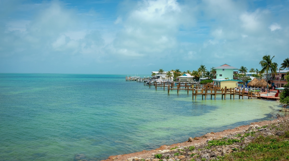  A view of Islamorada, Florida, characterized by an overcast sky with clouds and calm waters. Numerous small wooden docks line the shore, with no visible boats present. Small houses of various colors are dispersed across the landscape.