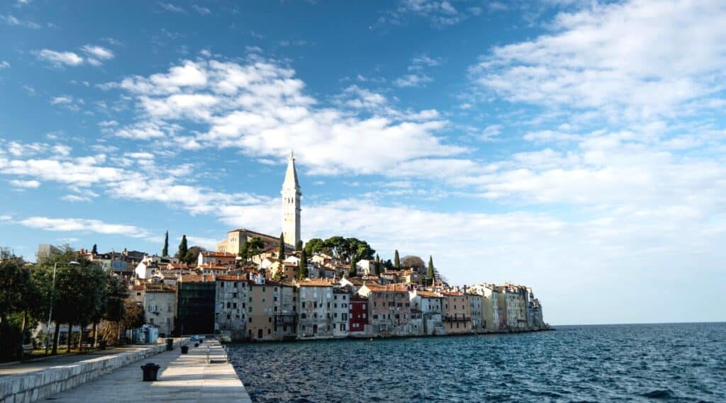 A view of the Croatian city of Rovinj in the region of Istria. The image captures a tranquil scene with a calm sea, an absence of people, and a few clouds in the sky. The cityscape comprises houses of varied colors, each with brown roofs.