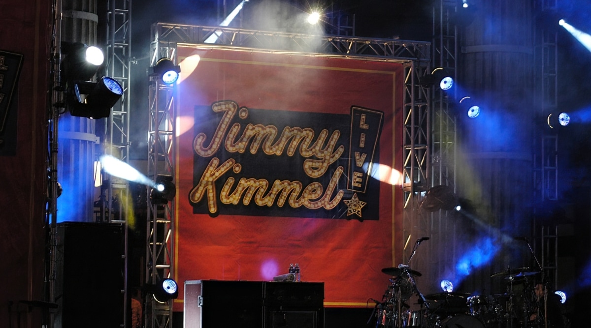 A photograph capturing the backstage area of Jimmy Kimmel Live with prominent signage displaying the show's name. The image features an array of spotlights surrounding the banner.