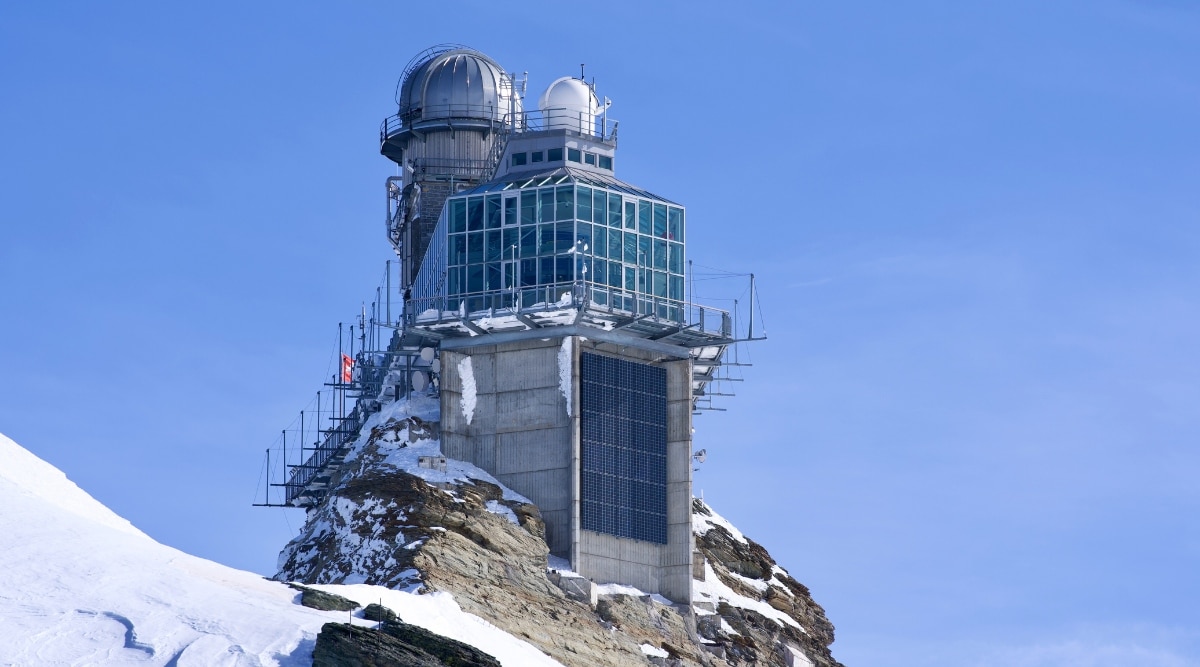 A photograph depicting Jungfraujoch Railway Station in Switzerland. The image offers a clear view of the station's infrastructure and surrounding landscape. Situated against a backdrop of mountainous terrain.