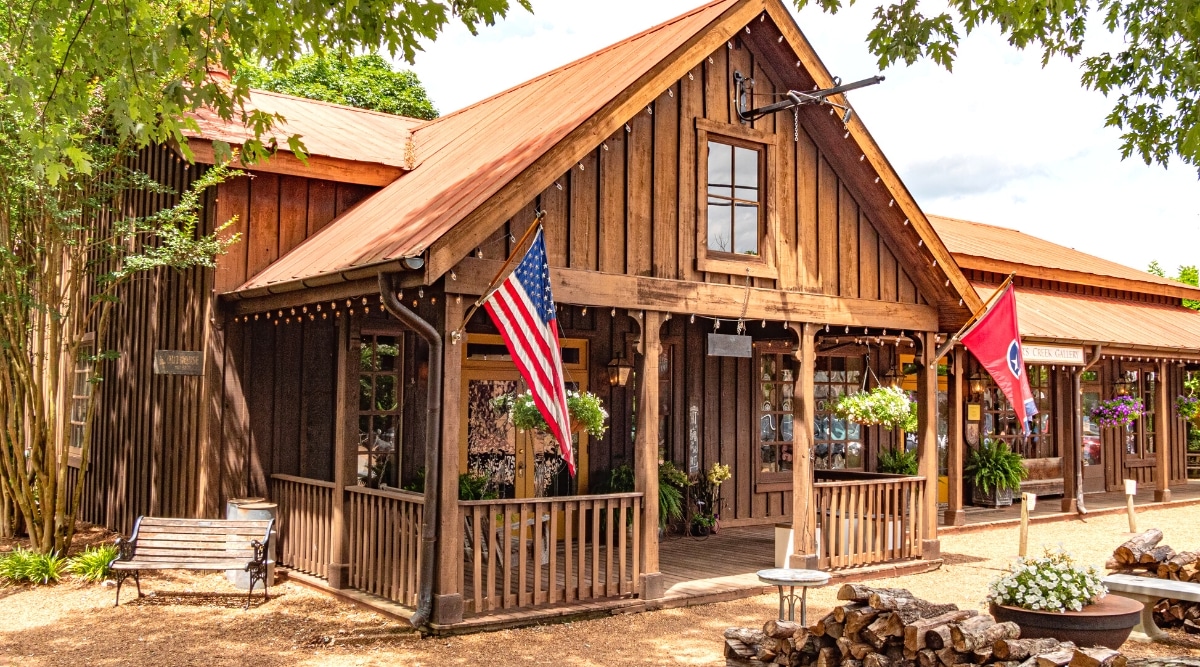 An exterior view of Leiper's Creek Gallery in Tennessee, constructed with brown wood tones. The image features the entrance adorned with two flags, one representing the USA and the other the state of Tennessee. A bench is situated on the side. The architectural elements of the gallery's exterior are characterized by a wooden construction.
