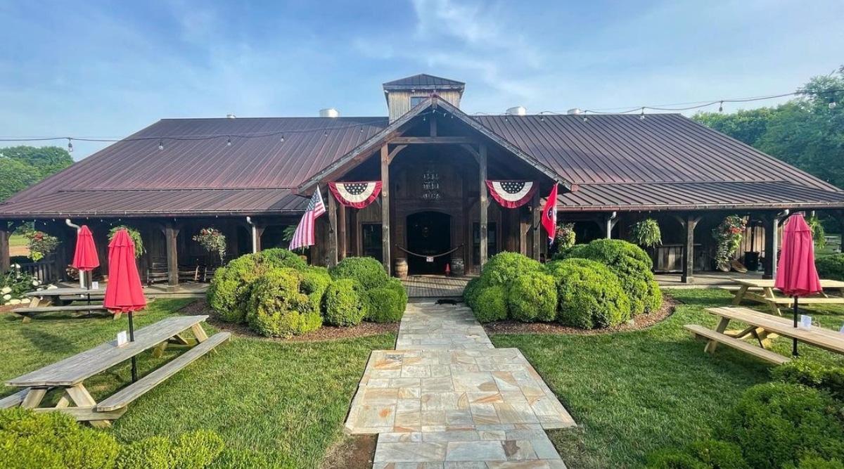 A direct view of Leipers Fork Distillery, prominently featuring the entrance with flags on both sides— the American flag and the Tennessee state flag. The architectural details of the distillery are visible, and rounded bushes are present on both sides of the entrance. Benches and folded red umbrellas provide seating arrangements in the immediate vicinity.