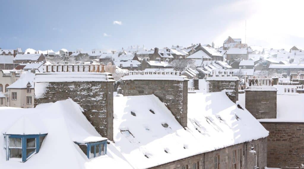 A view capturing snow-covered roofs of houses in Lerwick, Scotland. The image displays the residential area under a blanket of snow, with houses featuring varied architectural styles. The weather conditions are characterized by a layer of snow on rooftops.