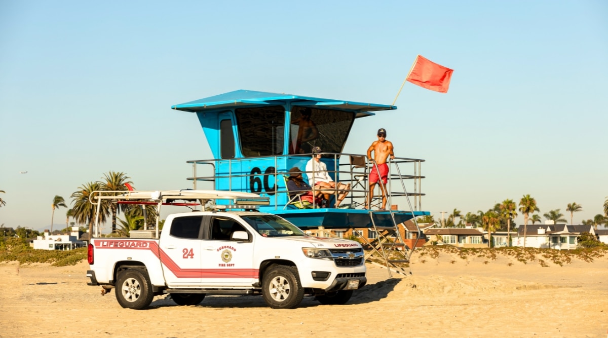 The image captures a lifeguard station on Coronado Beach, featuring a blue birch-colored structure marked with the number 60. A rescue vehicle is present near the station, and three lifeguards are observed in the vicinity. The sky overhead is clear, devoid of any clouds.