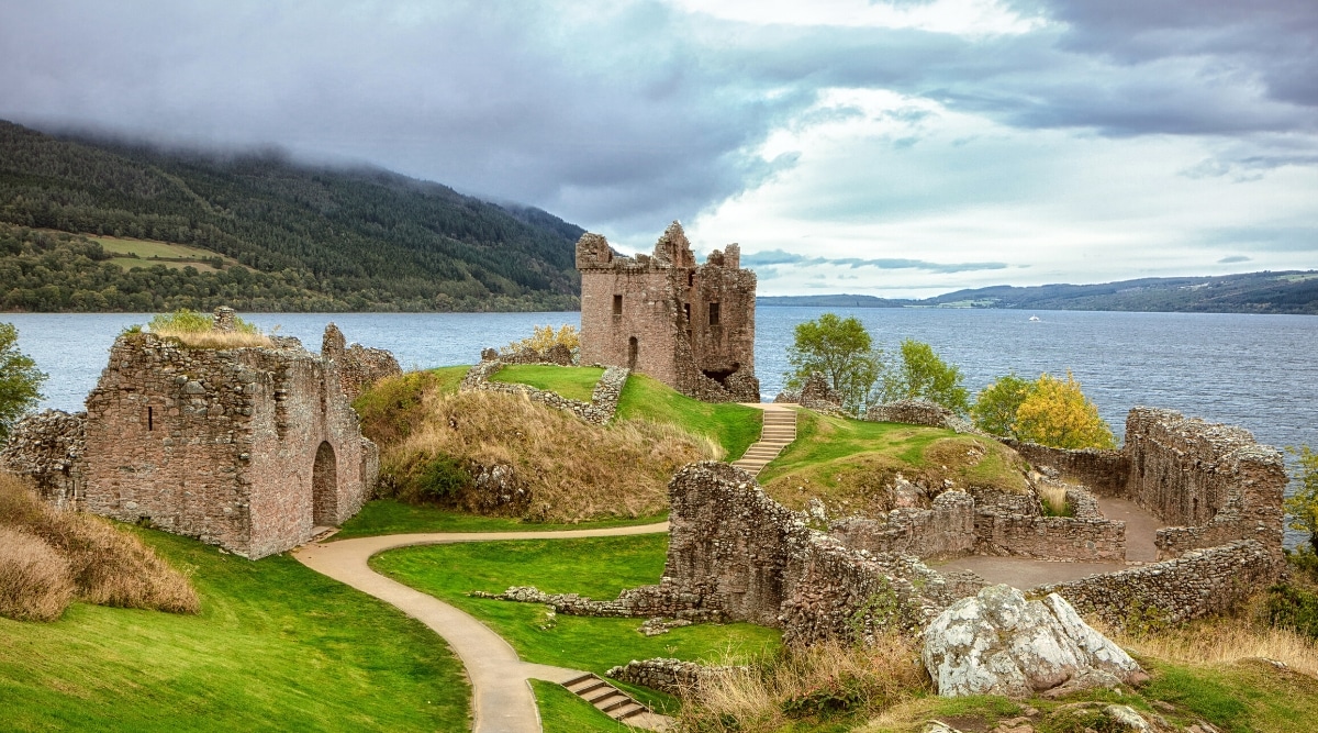 A photograph depicting Urquhart Castle situated on the shores of Loch Ness in Scotland. The image features the castle's ruins against the backdrop of the expansive lake. The surrounding natural environment consists of the lake and distant hills.