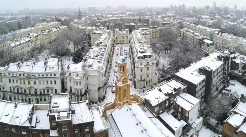 An aerial view capturing the snowy landscape of Notting Hill in London, United Kingdom. The image features the residential and commercial areas of Notting Hill covered in snow, revealing a winter scene. The rooftops, streets, and open spaces are dusted with snow.
