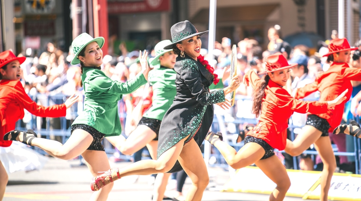 The image depicts a street performance featuring girls in red, green, and black costumes with hats, engaged in a dance routine. The dancers are showcased against the backdrop of a street, with numerous spectators observing the performance in the background. 
