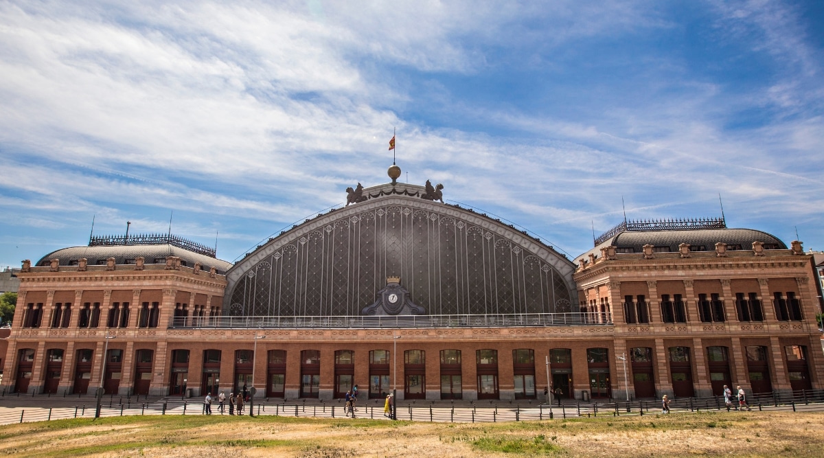 A photograph capturing Madrid Puerta de Atocha, a prominent train station in the heart of Madrid. The image displays the station's architectural features, including the main building. Commuters and travelers are visible around the station.