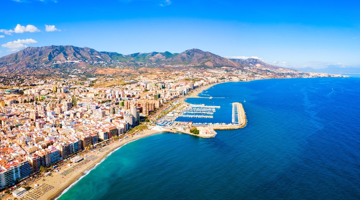 A captivating aerial perspective capturing the scenic beauty of Malaga, Spain. The image reveals a stunning contrast between the azure sea on the right and the vibrant cityscape on the left. In the center, a prominent pier showcases moored ships. Towering in the background, rocky mountains.