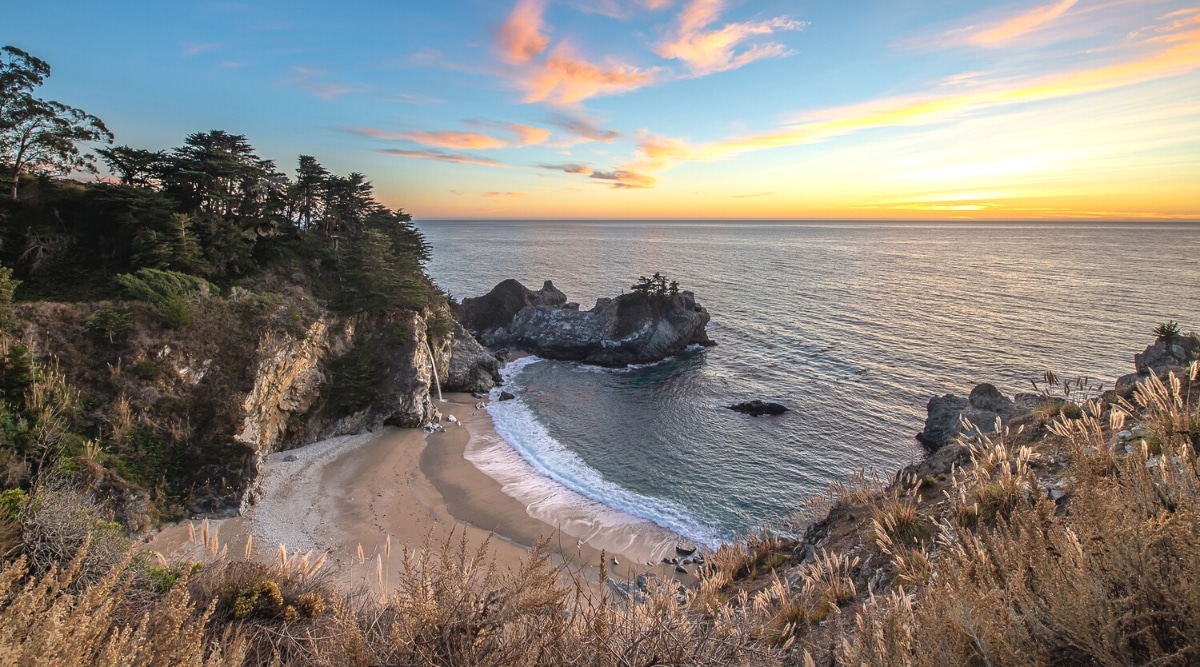 An aerial view capturing McWay Beach in Big Sur, characterized by small rocks, abundant greenery, and the Pacific Ocean. The image depicts the coastal landscape during sunset, featuring a serene combination of rocky formations and lush vegetation.