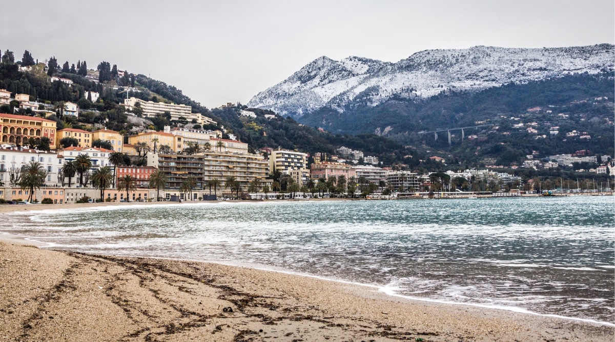 A photograph depicting the beach in Menton, France, set against the city backdrop. The image showcases the sandy shore and coastline with minimal foreground activity. In the background, the cityscape of Menton is visible.