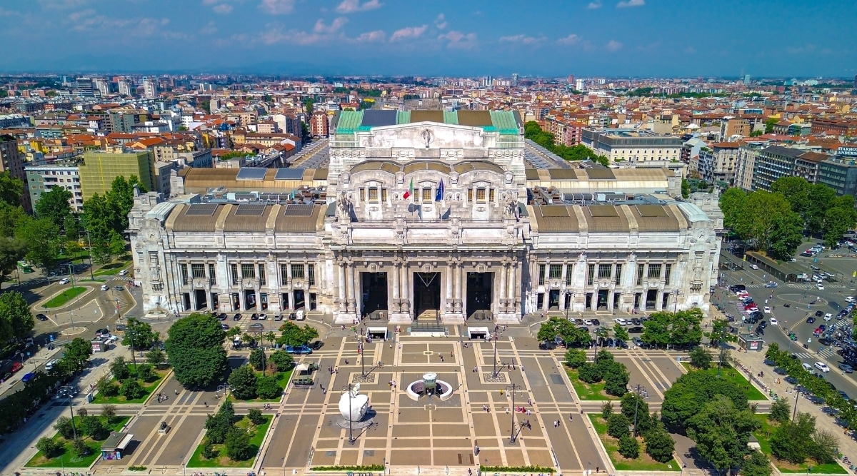 A photograph capturing Milano Centrale, the main railway station in Milan. The image features the exterior of the station, showcasing its architectural details. Commuters and travelers are visible in the foreground.The station's exterior, characterized by classical architectural elements.
