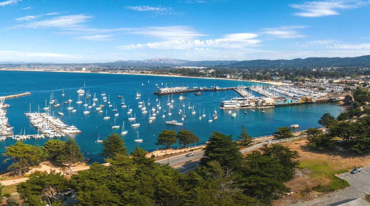 An aerial view capturing Monterey town in California, featuring numerous yachts docked by the pier. The image provides a comprehensive overview of the coastal town's layout, emphasizing the presence of yachts along the waterfront.