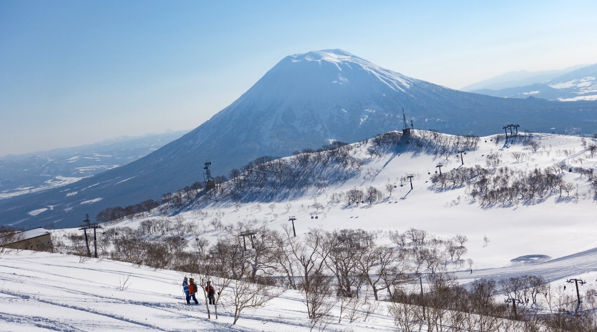 A view of snow-capped hills at Niseko United Ski Resort. The image depicts a winter landscape with minimal human presence, as only three individuals are observed standing in the distance. The snow-covered hills provide a serene backdrop, showcasing the resort's natural surroundings during the winter season.