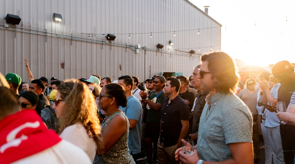 A photograph capturing a crowd gathered in an open-air space, facing a stage, with a large metal hangar in the background.