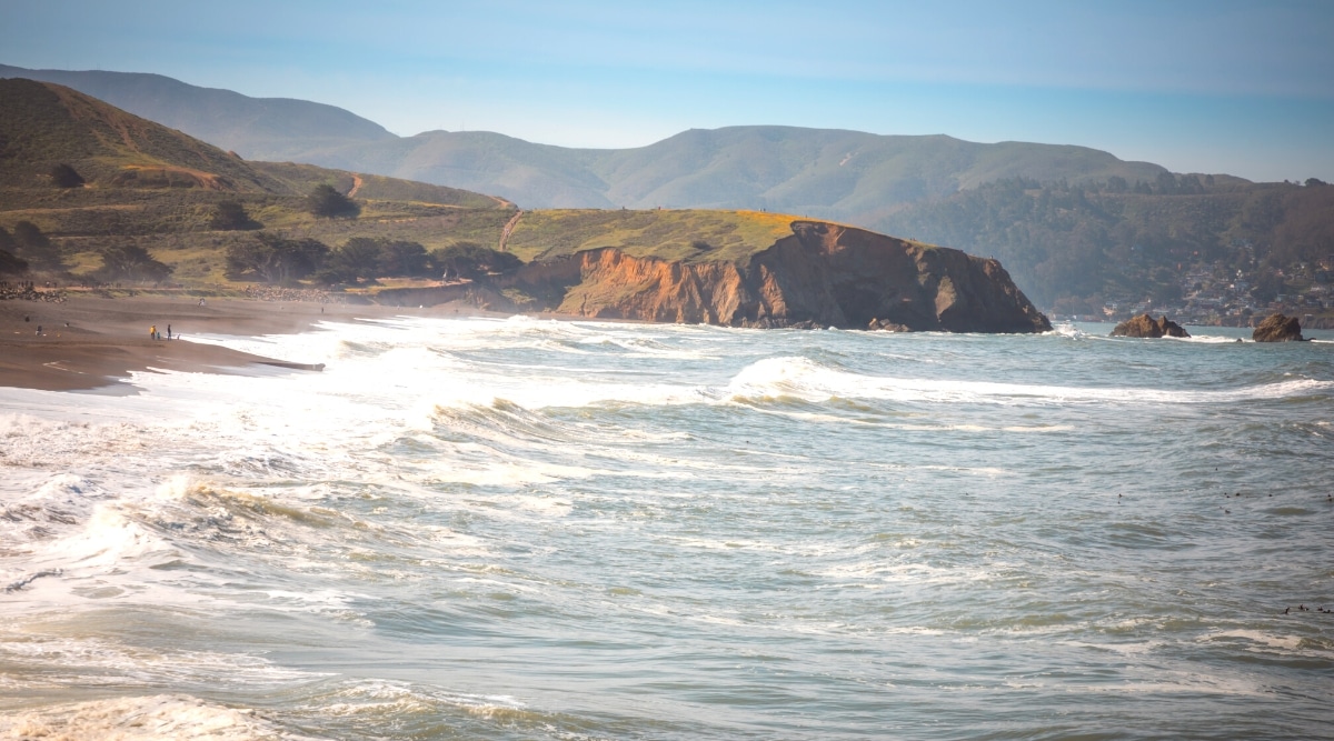 A straightforward photograph capturing Pacifica State Beach, with the beach visible on the left and the ocean on the right. In the background, the scene extends to showcase mountains and hills.