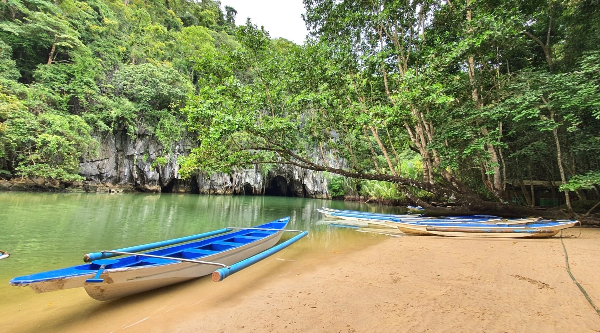 The photograph depicts a scene at Puerto Princesa Subterranean River National Park in Palawan, Philippines. Several small blue boats are anchored near the shore, with no visible people.