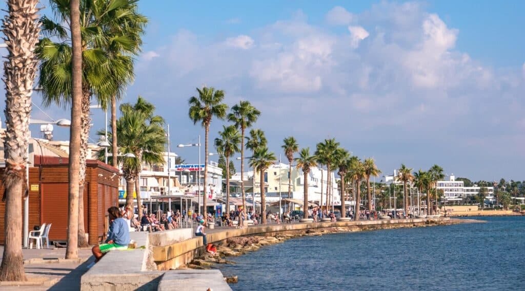 The photograph captures the Paphos embankment or promenade in Cyprus, featuring palms, people, shops, restaurants, and the blue Mediterranean Sea. The scene portrays a typical coastal setting of one of Cyprus's renowned resorts. Palm trees line the promenade.