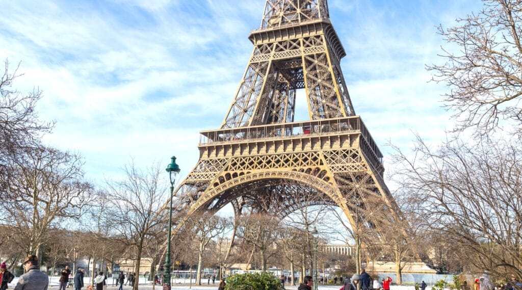 A close-up depiction of half of the Eiffel Tower during the winter season. The scene includes a snow-covered street and people walking in the square beneath the Eiffel Tower. The architectural details of the tower's structure are visible, with a neutral winter sky in the background.