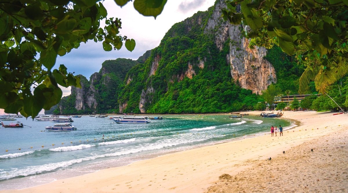 A visual depiction of Phuket Beach showcasing the natural elements of sand, greenery, rocks, and the ocean. The image features boats anchored along the shoreline and several people standing on the beach. The sandy expanse is framed by coastal rocks.