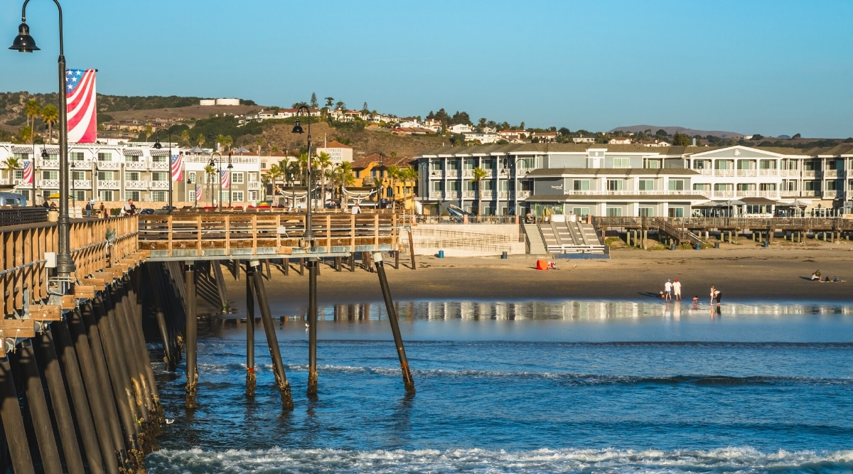 A photograph capturing the coastal scene of Pismo Beach, California, featuring the renowned pier, surrounding hills, buildings, and individuals on the beach.