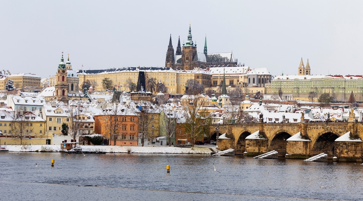 The image captures a winter scene of Snowy Prague Lesser Town, featuring Prague Castle and Charles Bridge above the River Vltava in the Czech Republic.