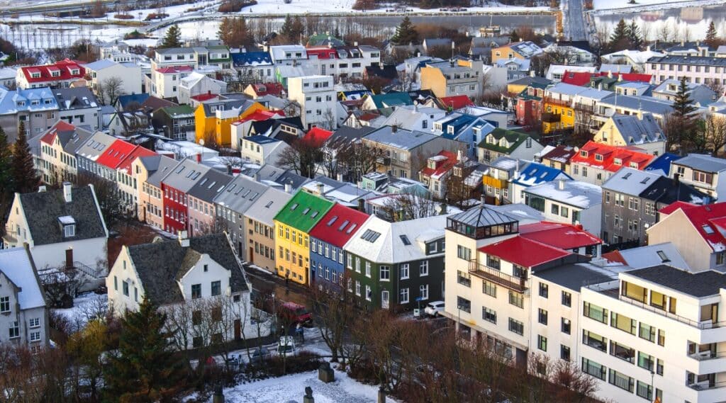 A bird's eye view capturing the cityscape of Reykjavik during winter. The image showcases the diverse rooftops and houses adorned in various colors, including red, green, black, white, gray, yellow, and blue. The streets are covered with snow, while the roofs appear to be mostly devoid of snow.The architectural diversity is evident in the range of colors and styles of the buildings, contributing to the city's visual landscape.