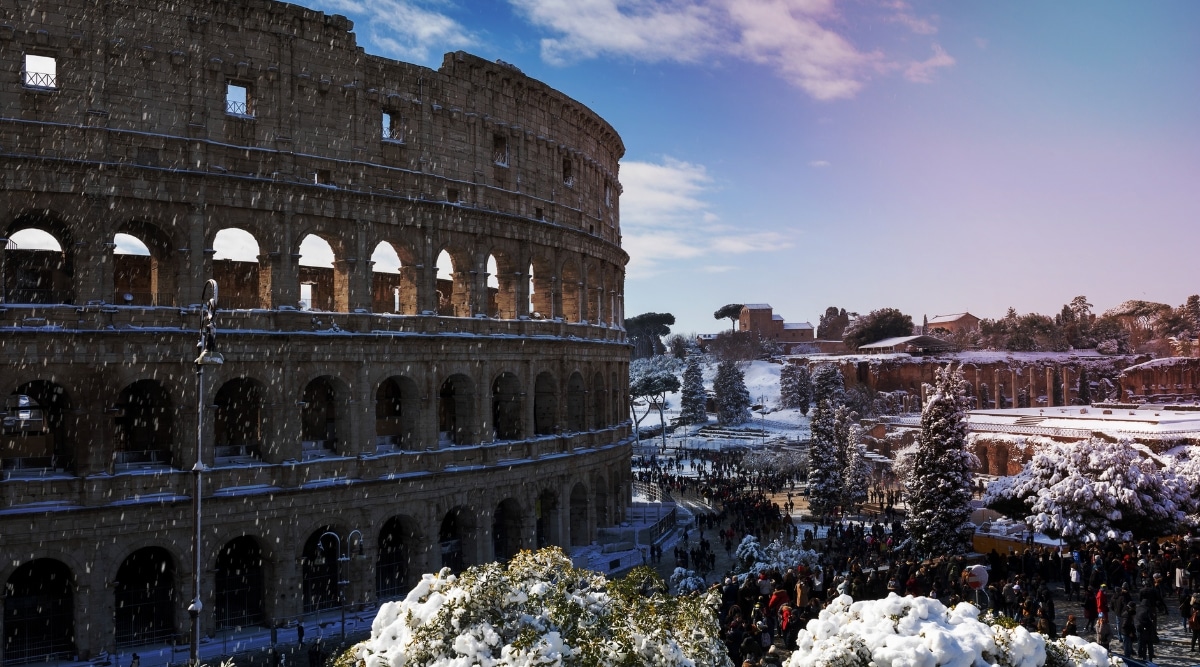 A view capturing the Colosseum covered in snow, with bright weather conditions and snowfall. The iconic structure is surrounded by snow-laden trees along the street, presenting a winter scene. 