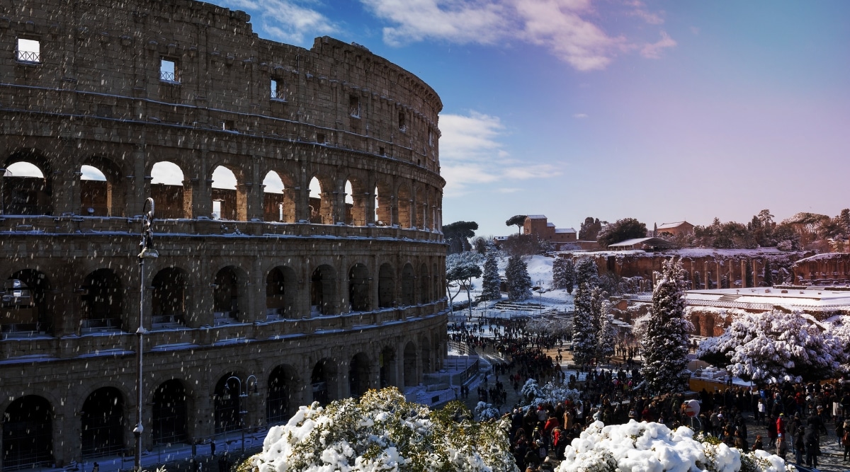 A view capturing the Colosseum covered in snow, with bright weather conditions and snowfall. The iconic structure is surrounded by snow-laden trees along the street, presenting a winter scene.